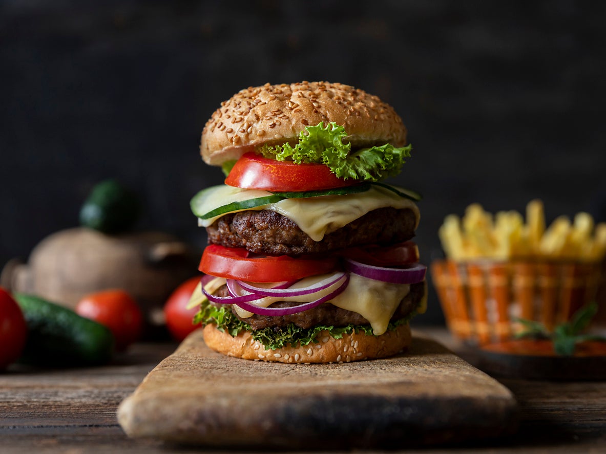 Hamburger with tomatoes, onions, cucumber, lettuce and melting cheese served on a rustic wooden board with french fries and vegetables in background
