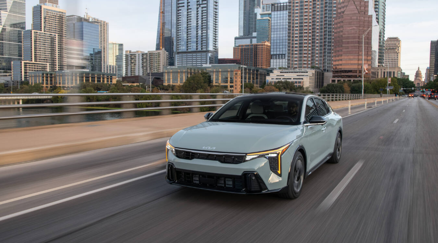 Front three-quarter view of a light sage green 2027 Kia K4 compact sedan driving across a city bridge with skyscraper skyline in the background, showcasing its vertical LED headlights and bold fastback design for drivers in Waukegan, IL, available at Classic Kia.