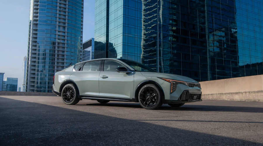 Side profile of a light sage green 2027 Kia K4 sedan parked against modern glass high-rise buildings, highlighting its fastback roofline, vertical LED DRLs, and black alloy wheels for commuters in Waukegan, IL, available at Classic Kia.