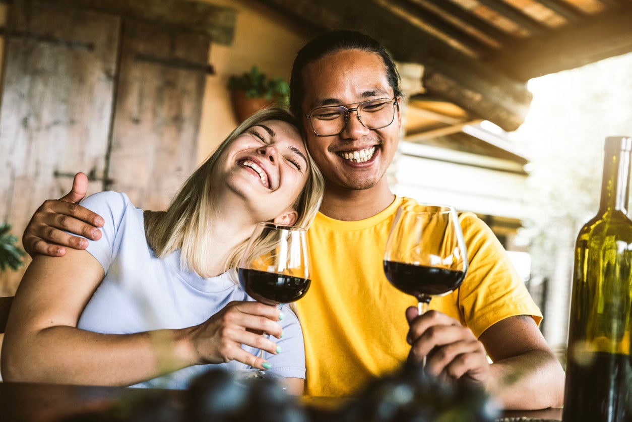 Young couple in love on a date having fun drinking red wine in farmhouse garden - Multiracial boyfriend and girlfriend enjoying wine tasting weekend experience at restaurant patio - Focus on guy face