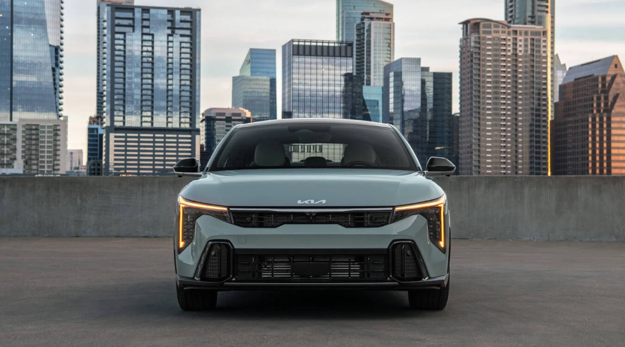 Direct front-facing view of a light sage green 2027 Kia K4 sedan highlighting its illuminated amber vertical LED daytime running lights, wide front grille, and Kia hood badge with a city skyline in the background, available at Classic Kia in Waukegan, IL.