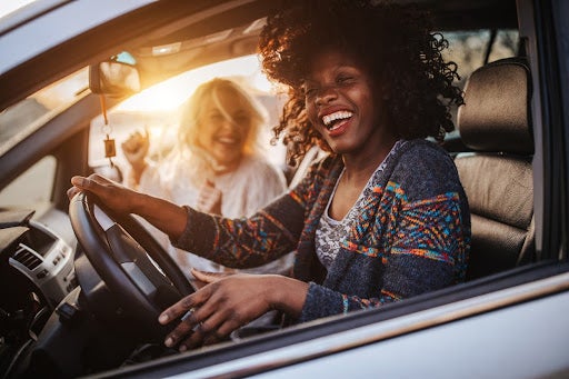 Two young cheerful female friends having fun on road trip in car at sunset