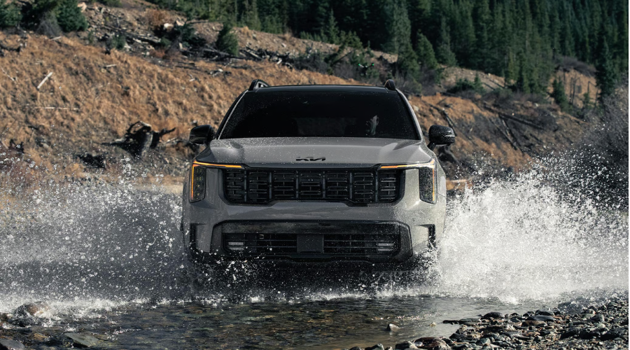 Front view of a grey 2026 Kia Sorento SUV driving head-on through a shallow riverbed, splashing water dramatically with amber LED daytime running lights illuminated, showcasing all-weather AWD performance for fall camping near Illinois Beach State Park in Zion and Lake County, IL.