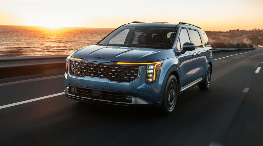 Dynamic front-three-quarter view of a metallic blue 2027 Kia Carnival cruising along a coastal highway at sunset, highlighting 3.5L V6 engine performance available at Classic Kia near Zion and Waukegan, IL.