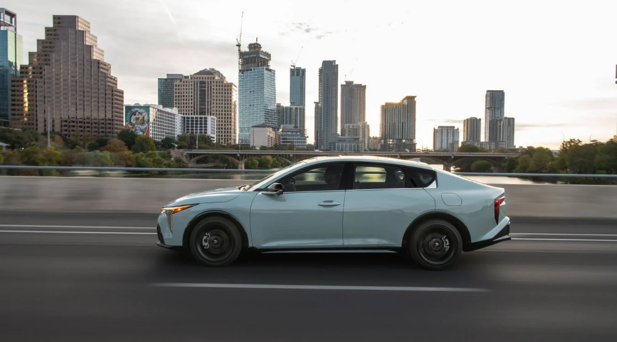 Full side profile of a light sage green 2027 Kia K4 sedan driving in motion across a multi-lane highway bridge with a city skyline in the background, highlighting its long wheelbase and aerodynamic fastback silhouette for daily commuters in Waukegan, IL, available at Classic Kia.