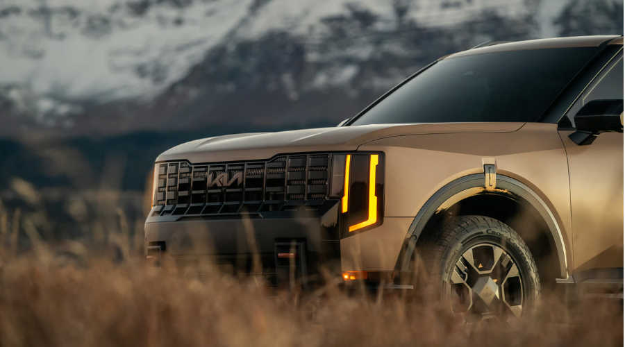 Close-up front three-quarters view of a bronze 2026 Kia Telluride X-Pro featuring amber vertical LED daytime running lights, a bold dark front grille with Kia badge, and Continental tires parked in tall golden autumn grass, illustrating rugged capability for fall camping trips in Waukegan and Lake County IL.