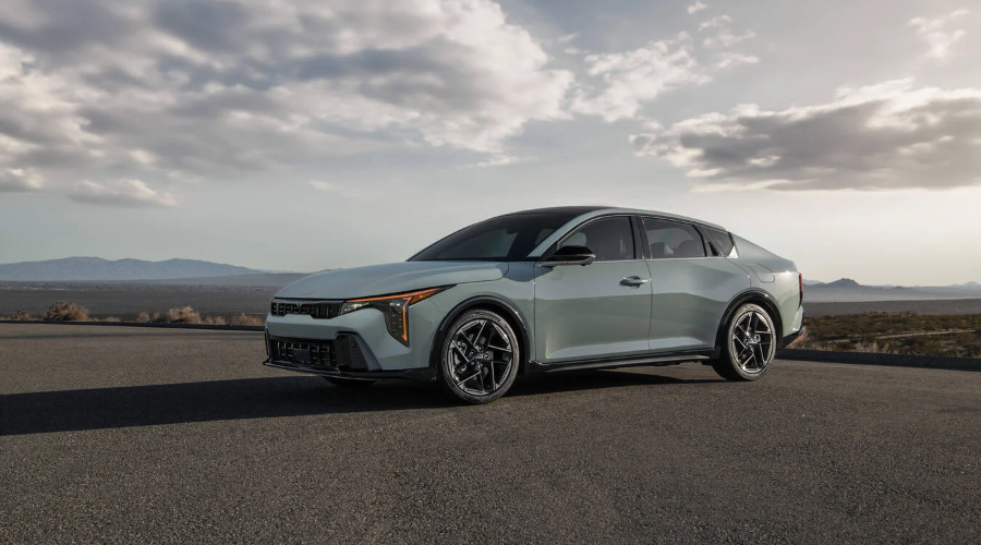 Front three-quarter view of a light sage green 2027 Kia K4 sedan parked on an open road under a cloudy sky, showcasing its geometric alloy wheels, vertical LED lights, and fastback styling for drivers in Waukegan, IL, available at Classic Kia.