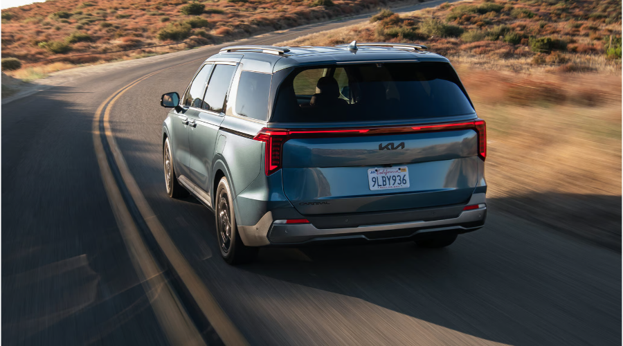 Rear three-quarter driving view of a metallic blue 2027 Kia Carnival navigating a curved highway, highlighting full-width LED taillights and rear spoiler available at Classic Kia serving Waukegan and Antioch, IL.