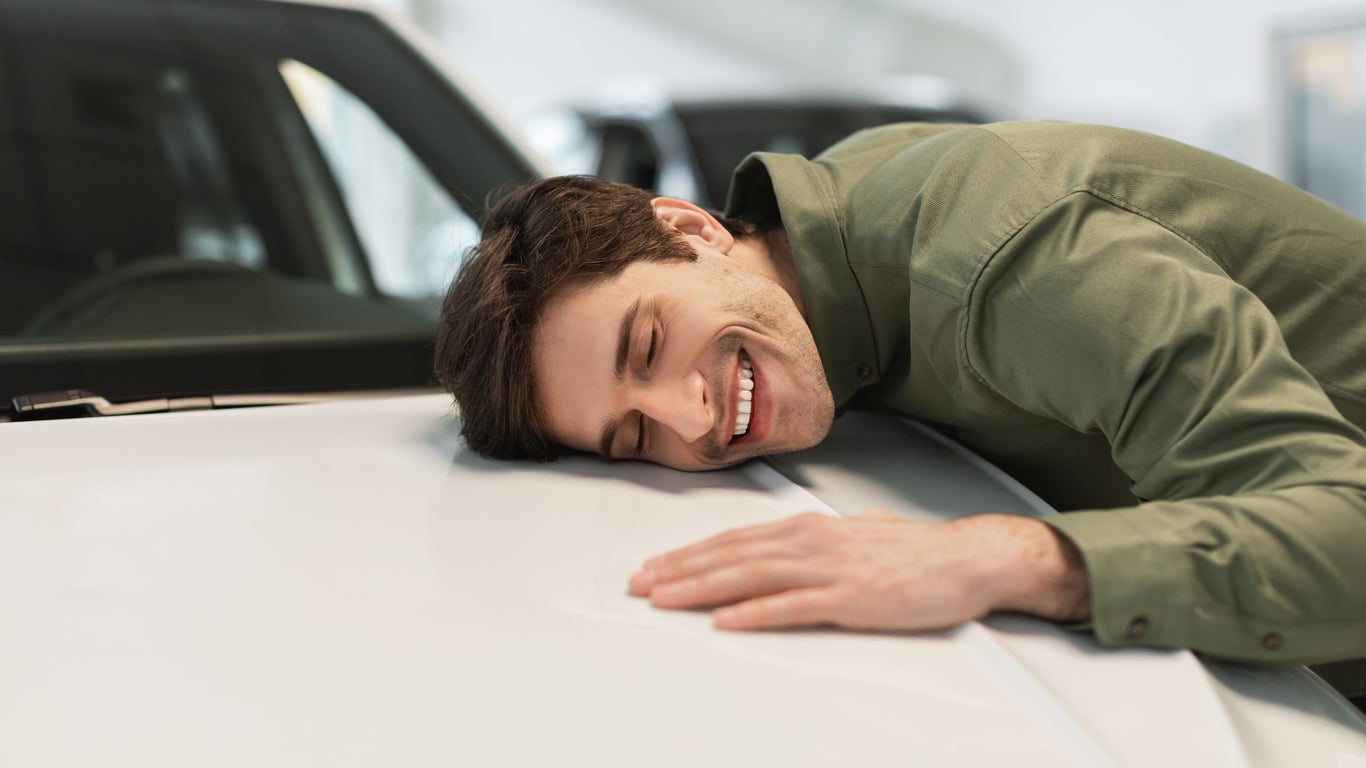 Happy young Caucasian guy hugging his brand new car, buying vehicle at dealership centre, panorama with copy space