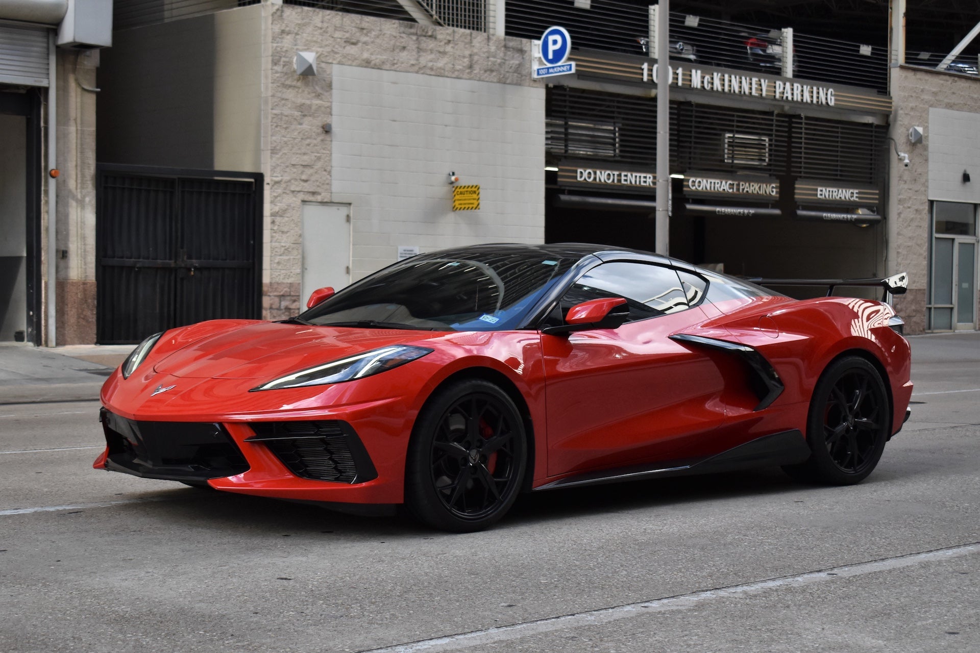 A portrait of a red and black Chevy Corvette C8 3LT sports car traveling near the McKenney Parking Garage in the downtown financial district of Houston.