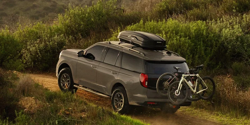 A Ford Expedition SUV driving on a dirt trail, equipped with a roof box and two bikes mounted on the rear.
