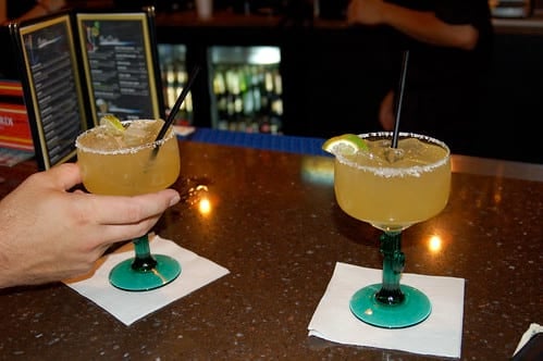Two margaritas on bar with straws and someone reaching to pick one up