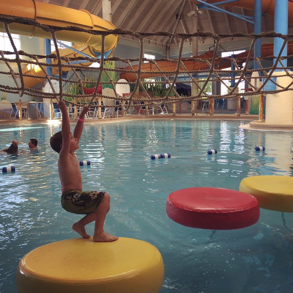 Kid climbing across a rope latter over an indoor pool