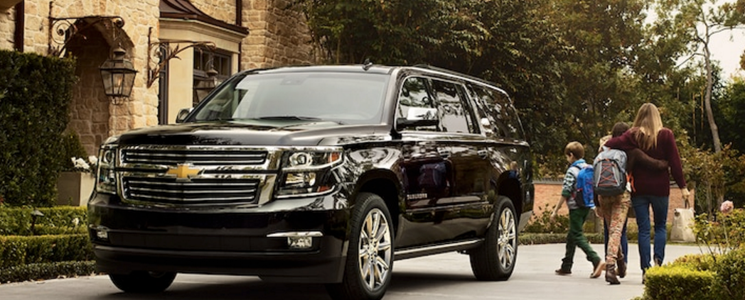 A black 2017 Chevy Suburban parked in front of the house. A woman with two kids carrying backpacks walk next to the SUV.