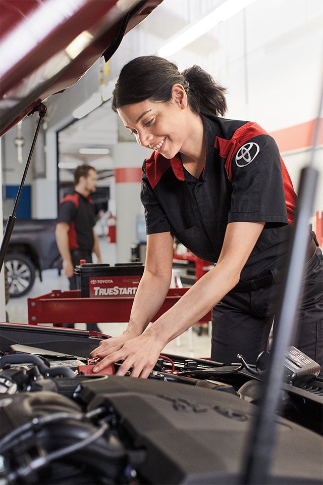 Everything You Need to Know about Your Car Battery at Bennett Toyota | Female Toyota Employee Under Hood of Car Checking Battery