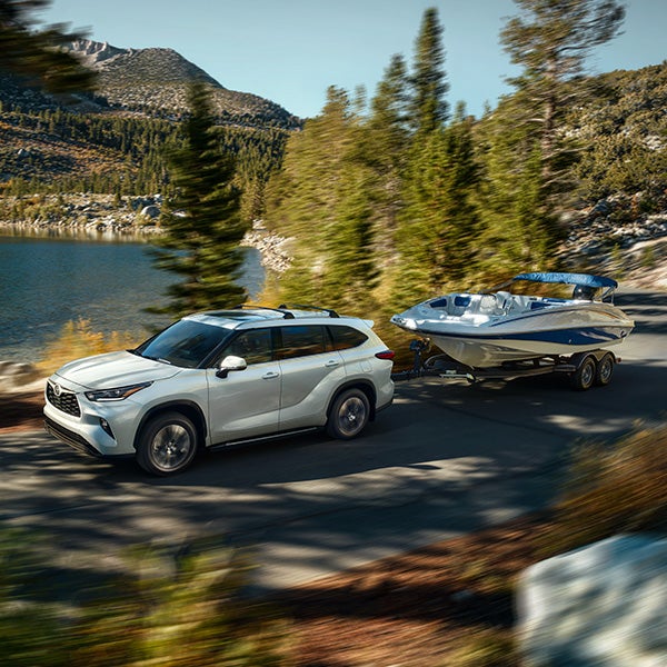 The 2022 Toyota Highlander vs. The 2021 Toyota Highlander at Bennett Toyota | White 2022 Toyota Highlander Driving By Lake While Towing a Boat Behind it