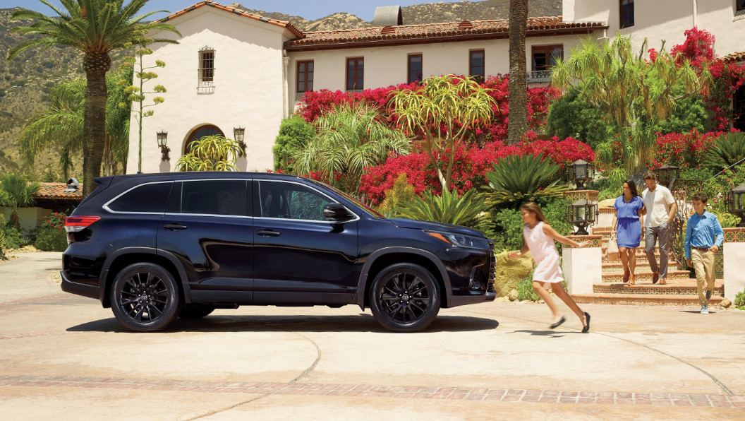 Image of a dark blue 2019 Toyota Highlander parked in a driveway with a family running up to it.